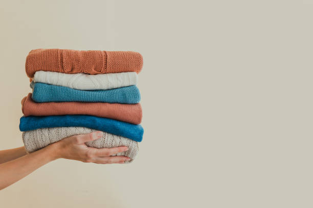 Stack of neatly folded sweaters of different colors held in hands against a plain background.