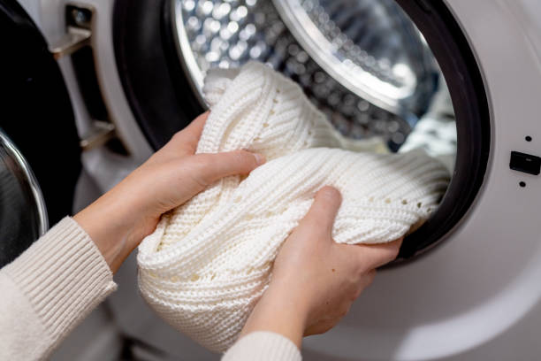 Hands placing a clean white sweater into a washing machine for proper laundry care.